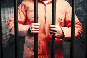 A prisoner in an orange jumpsuit holds prison bars.