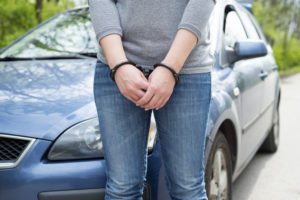 A woman handcuffed in front of a blue car.
